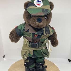 Puede incluir: Un oso de peluche marrón con uniforme y gorra de camuflaje con un parche circular. El uniforme incluye chaqueta, pantalón y cinturón. El oso también lleva botas negras. Las palabras "U.S. ARMY" son visibles en el uniforme.