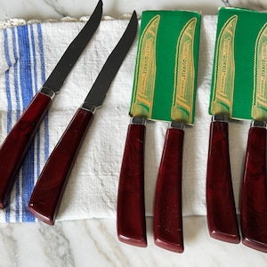 May include: Six stainless steel knives with dark red marbled handles. Two knives are unsheathed, while four are partially covered by green cardboard sleeves labeled "Quikit Quikkle Stainless". The knives feature serrated blades and curved handles.