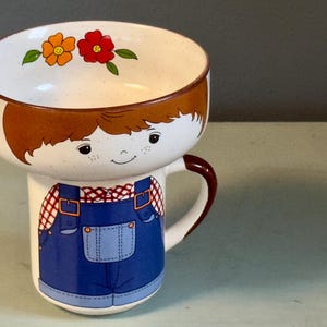 May include: A whimsical ceramic mug and bowl set. The bowl is decorated with orange and red flowers. The mug features a smiling boy with freckles wearing blue overalls. The handle is brown.