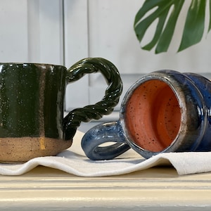 May include: Two handmade ceramic mugs. One is dark green with a twisted handle and a speckled finish. The other mug is blue with a ribbed design and a brown base. Both mugs have a rustic, artisanal aesthetic.