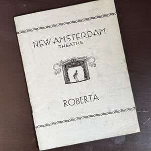 May include: A vintage theater program for "Roberta" from the New Amsterdam Theatre. The cover is off-white with black text and a decorative border. The program features a small illustration of a stage with a performer.