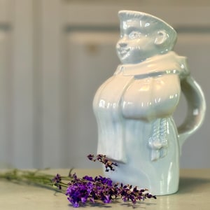 May include: A light blue ceramic pitcher in the shape of a monk, with a handle and a smiling face. A sprig of purple lavender rests in front of the pitcher.