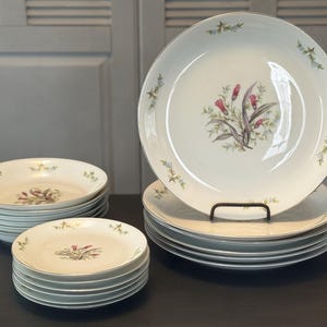 May include: A collection of white ceramic plates with a floral design. The plates have a gold rim and a central image of pink and purple flowers. The set includes dinner plates, salad plates, and bowls, stacked neatly on a dark surface.