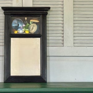 May include: Black framed mirror with a painted still life scene above the mirror. The painting includes a scale, lemons, and a metal bowl. The mirror is rectangular and the frame has a grooved design.
