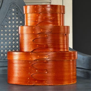 May include: A set of three oval-shaped wooden boxes stacked vertically, featuring a warm orange-brown finish. Each box has a lid with distinctive finger-jointed seams secured by small metal fasteners, creating a uniform appearance across the set.