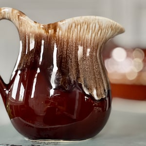 May include: A ceramic pitcher with a glossy, dark brown base and a lighter brown and white textured top. The pitcher has a curved handle and a spout for pouring. The design is reminiscent of vintage kitchenware.