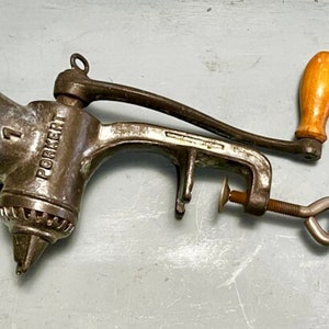 May include: An antique Porkert meat grinder, made of metal, with a wooden handle. The grinder is clamped to a surface and features the number "1" on the side. The handle is light brown.