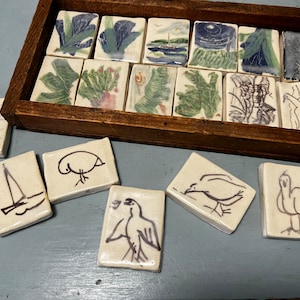 May include: A collection of small, rectangular ceramic tiles with hand-painted designs. The tiles feature various nature-inspired illustrations, including birds, sailboats, and foliage, in shades of blue, green, and black. The tiles are arranged in a wooden tray.