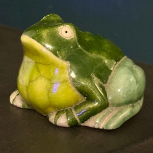 May include: A ceramic frog figurine in shades of green and yellow. The frog has a glossy finish and is sitting on a dark surface. The frog's belly is yellow with a cracked pattern, and the back is green.