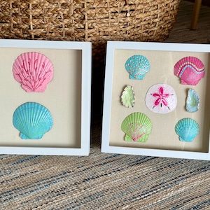 May include: Two square white frames containing collections of painted seashells. The left frame holds two large scallop shells in pink and blue. The right frame displays six smaller shells and a sand dollar, featuring pink, blue, and green colors.
