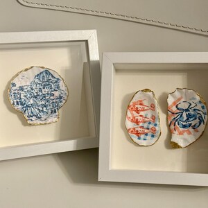 May include: Two square white shadow box frames containing decorative oyster shells. The left frame holds a shell painted with a blue and white coastal village scene. The right frame holds two shells, one with orange fish and one with a blue crab.