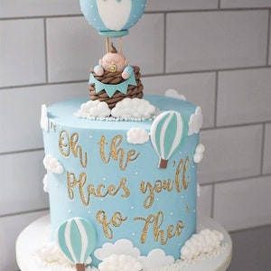 May include: A blue and white baby shower cake with a hot air balloon topper and the words "Oh the places you'll go Theo" written in gold glitter.