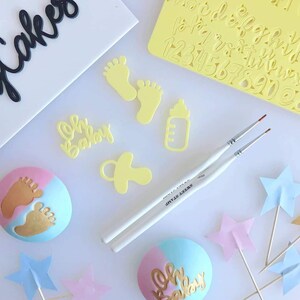 Könnte beinhalten: Ein Set gelber Fondant-Ausstecher zum Dekorieren von Kuchen und Cupcakes. Die Ausstecher beinhalten eine Babyflasche, einen Schnuller, zwei Fußabdrücke und die Worte "Baby". Zwei weiße Pinsel sind ebenfalls enthalten.