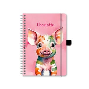 Könnte beinhalten: Rosa gebundenes Notizbuch mit einer Aquarell-Illustration eines bunten Ferkels. Das Notizbuch hat einen schwarzen Gummizugverschluss und der Name "Charlotte" ist auf dem Einband aufgedruckt.