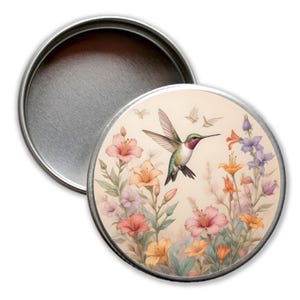 Könnte beinhalten: Eine runde Metalldose mit einem Kolibri im Flug, umgeben von bunten Blumen. Die Dose hat einen cremefarbenen Hintergrund mit einem hellbraunen Waschgang.