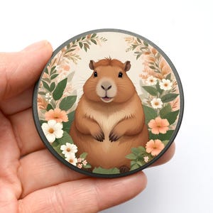 Könnte beinhalten: Runder Untersetzer mit einem Cartoon-Capybara, umgeben von Blumen. Das Capybara ist braun mit hellem Bauch und lächelt. Die Blumen sind weiß und pfirsichfarben, vor hellem Hintergrund.