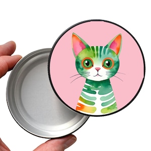Könnte beinhalten: Eine runde Metalldose mit Deckel. Der Deckel zeigt eine Aquarellillustration einer grünen Katze mit orangefarbenen und rosa Akzenten auf rosa Hintergrund. Die Dose ist geöffnet und zeigt das silberne Innere.