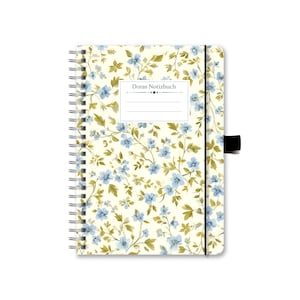 Könnte beinhalten: Ein weißes Spiralnotizbuch mit einem Blumenmuster mit blauen Blüten und grünen Blättern. Der Umschlag hat ein Etikett mit der Aufschrift "Doras Notizbuch".