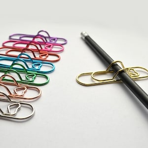 Könnte beinhalten: Eine Sammlung von Metall-Büroklammern in verschiedenen Farben wie Lila, Pink, Rot, Blau, Grün, Kupfer und Silber, die in einer vertikalen Reihe angeordnet sind. Eine einzelne goldene Büroklammer ist an einem schwarzen Stift rechts daneben befestigt.