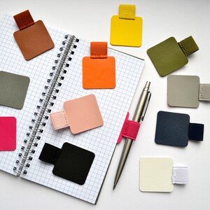 Könnte beinhalten: Eine Sammlung quadratischer Stifthalter mit elastischen Schlaufen in verschiedenen Farben wie Braun, Gelb, Olivgrün, Orange, Hellrosa, Schwarz, Grau, Marineblau und Creme, angeordnet um ein spiralgebundenes Notizbuch mit Karomuster und einen silbernen Metallstift.