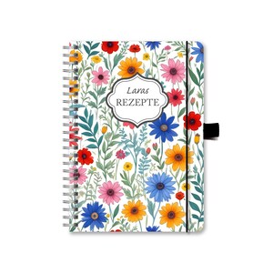 Könnte beinhalten: Ein weißes Spiralblock mit einem Blumenmuster. Der Einband zeigt eine Vielzahl von bunten Blumen, darunter rote, gelbe, blaue und rosa. Der Text "Laras REZEPTE" ist in Schwarz auf einem weißen Etikett gedruckt.