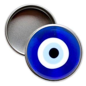 Könnte beinhalten: Eine kleine, runde, silberne Dose mit einem blauen bösen Auge auf dem Deckel. Das böse Auge ist ein Symbol für Schutz vor Pech und Neid.