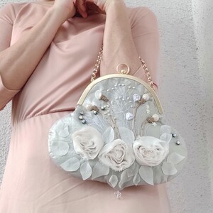 May include: A light gray, heart-shaped handbag with a gold chain strap. The bag is adorned with white fabric roses, delicate floral embroidery, and small, sparkling embellishments. The clasp is gold.