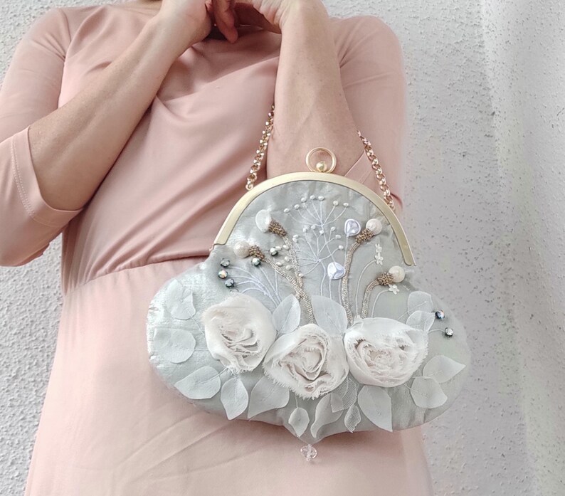 May include: A light gray, heart-shaped handbag with a gold chain strap. The bag is adorned with white fabric roses, delicate floral embroidery, and small, sparkling embellishments. The clasp is gold.
