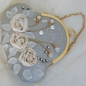 May include: A heart-shaped, light gray clutch with a gold chain strap. The clutch is embellished with three cream-colored fabric roses, white leaves, and beaded accents. The frame is gold-toned.