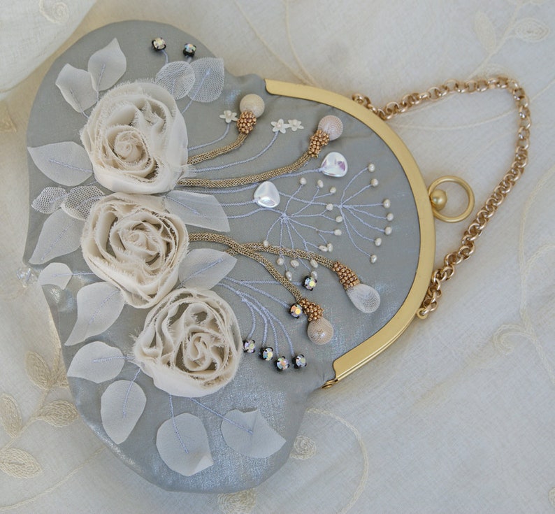 May include: A heart-shaped, light gray clutch with a gold chain strap. The clutch is embellished with three cream-colored fabric roses, white leaves, and beaded accents. The frame is gold-toned.