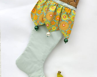 Puede incluir: Un calcetín navideño decorativo con un cuerpo verde claro, una parte superior acolchada marrón adornada con pedrería y una sección con volantes con un estampado floral en amarillo, naranja y azul. La punta es amarilla con detalles florales azules y una pequeña campana dorada.