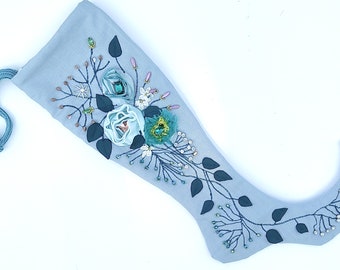 Puede incluir: Un calcetín navideño azul claro con adornos florales. Presenta bordados de vid, hojas azul oscuro y rosas de cinta azul claro. Una pequeña campana dorada cuelga de la punta. Una cuerda azul está unida en la parte superior.