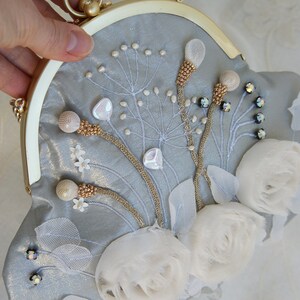 May include: A light blue clutch with a gold frame and clasp. The clutch is decorated with white floral embellishments, beaded accents, and embroidered details. The bag has a round handle.