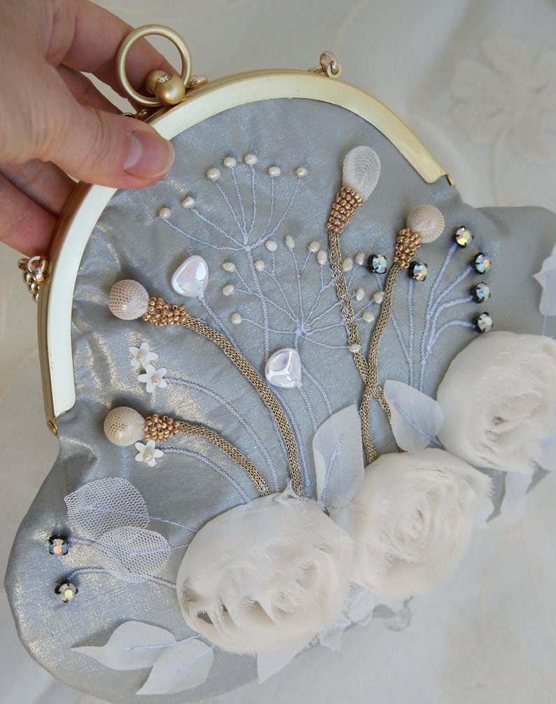 May include: A light blue clutch with a gold frame and clasp. The clutch is decorated with white floral embellishments, beaded accents, and embroidered details. The bag has a round handle.