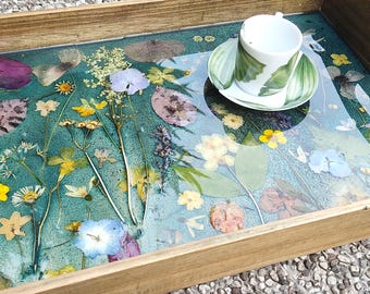 May include: A rectangular wooden serving tray featuring a teal resin base embedded with a variety of pressed dried flowers and leaves in shades of yellow, blue, purple, and green. A white ceramic teacup and saucer with green leaf patterns sits on top.