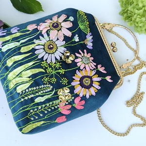 K&ouml;nnte beinhalten: Eine dunkelblaue Clutch aus Samt mit einer goldenen Kettenkette. Die Tasche ist mit gestickten Blumen in Rosa-, Lila- und Gelbt&ouml;nen verziert.