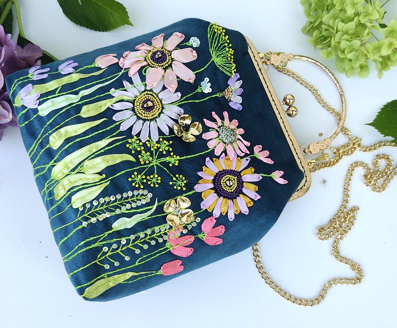 K&ouml;nnte beinhalten: Eine dunkelblaue Clutch aus Samt mit einer goldenen Kettenkette. Die Tasche ist mit gestickten Blumen in Rosa-, Lila- und Gelbt&ouml;nen verziert.