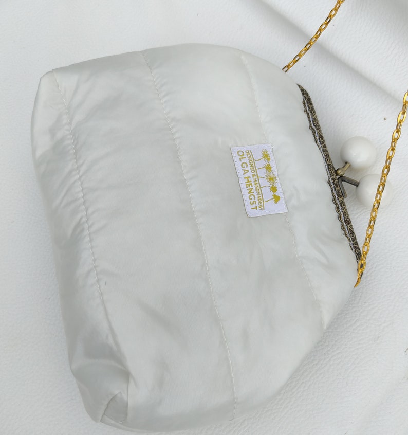 May include: White quilted clutch bag with a gold chain strap and a white label that reads "DESIGNED & HANDMADE BY OLGA HENGST".