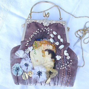 Puede incluir: Una cartera de mano de terciopelo malva con una correa de cadena dorada y un cierre de broche. La cartera presenta una imagen bordada de una madre y un hijo, rodeada de flores blancas y perlas.