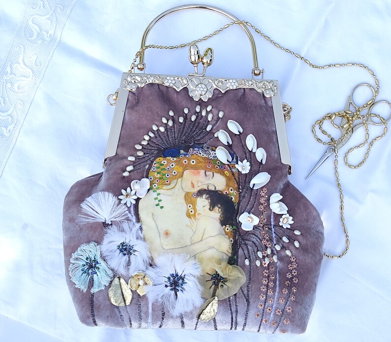 Puede incluir: Una cartera de mano de terciopelo malva con una correa de cadena dorada y un cierre de broche. La cartera presenta una imagen bordada de una madre y un hijo, rodeada de flores blancas y perlas.