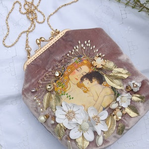 Puede incluir: Un bolso de mano de terciopelo malva con una correa de cadena dorada. El bolso presenta una pintura de una madre y un hijo con flores blancas y detalles dorados.