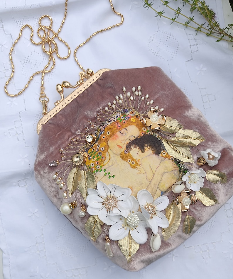 Puede incluir: Un bolso de mano de terciopelo malva con una correa de cadena dorada. El bolso presenta una pintura de una madre y un hijo con flores blancas y detalles dorados.
