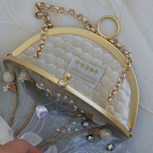 May include: A light blue clutch with a gold-tone frame and chain strap. The interior is quilted white fabric. Embellishments include beads, crystals, and floral accents. The label reads "OLGA HENGST".