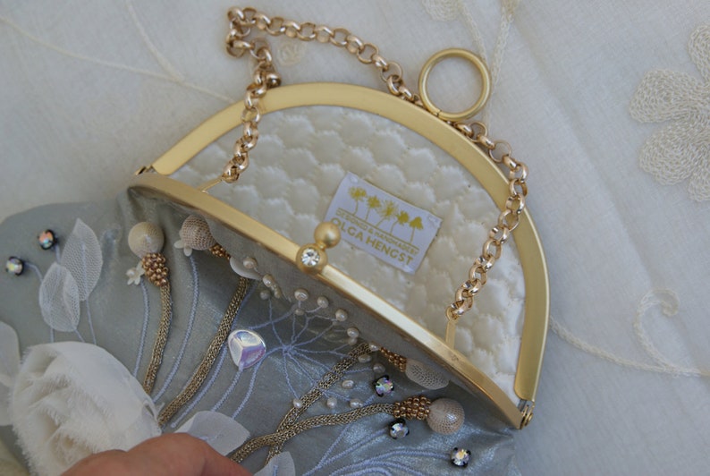 May include: A light blue clutch with a gold-tone frame and chain strap. The interior is quilted white fabric. Embellishments include beads, crystals, and floral accents. The label reads "OLGA HENGST".