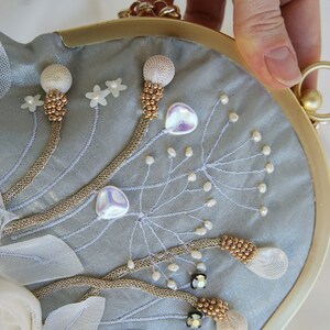 May include: A light blue satin clutch with gold-tone hardware and a chain strap. The clutch is embellished with beaded floral designs, white fabric flowers, and iridescent accents. The frame is a rounded shape.