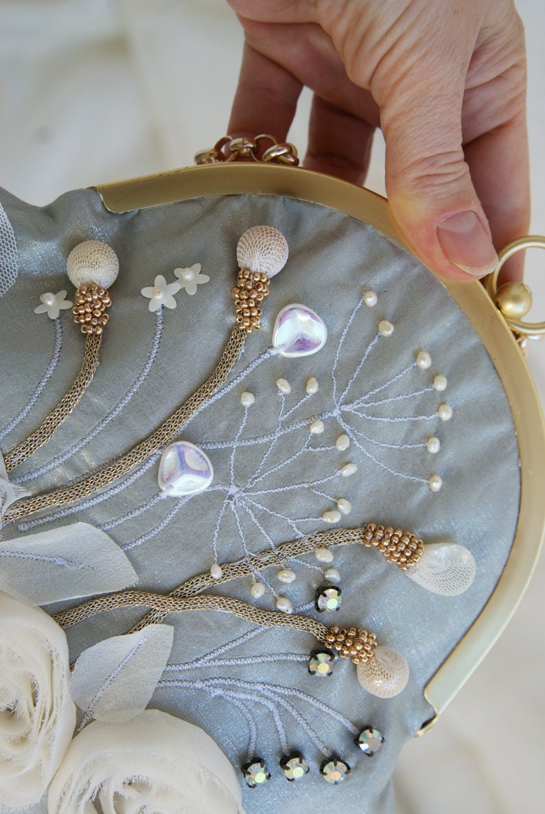May include: A light blue satin clutch with gold-tone hardware and a chain strap. The clutch is embellished with beaded floral designs, white fabric flowers, and iridescent accents. The frame is a rounded shape.