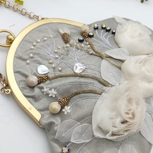 May include: A gray, heart-shaped handbag with gold-tone hardware and a chain strap. The bag is embellished with white floral accents, pearls, and beaded details. The design includes embroidered elements and iridescent gems.