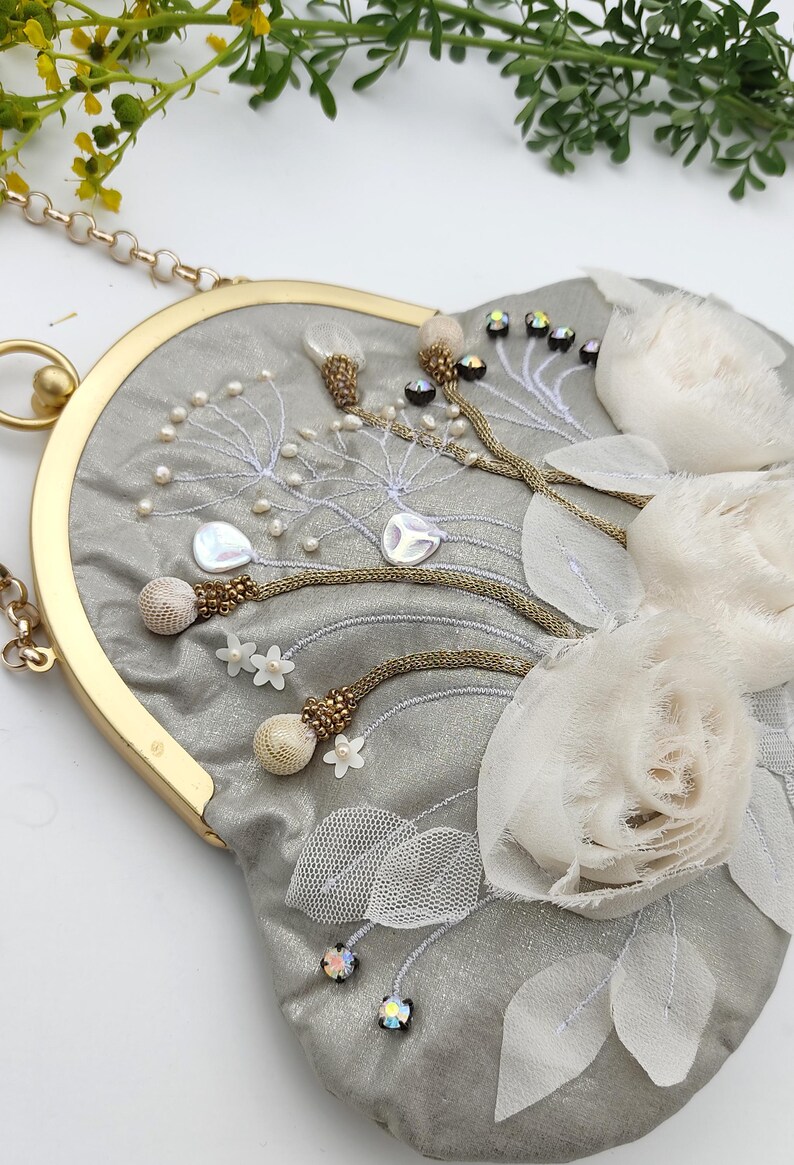 May include: A gray, heart-shaped handbag with gold-tone hardware and a chain strap. The bag is embellished with white floral accents, pearls, and beaded details. The design includes embroidered elements and iridescent gems.