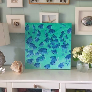 K&ouml;nnte beinhalten: Ein Gem&auml;lde, das einen Schwarm blauer Fische zeigt, die in einem t&uuml;rkisfarbenen und gr&uuml;nen Ozean schwimmen. Das Gem&auml;lde ist gerahmt und h&auml;ngt an einer wei&szlig;en Wand &uuml;ber einer wei&szlig;en Kommode.