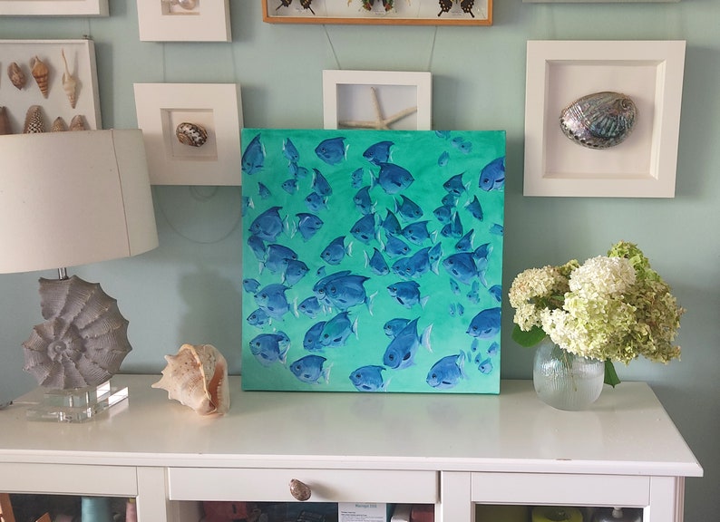 K&ouml;nnte beinhalten: Ein Gem&auml;lde, das einen Schwarm blauer Fische zeigt, die in einem t&uuml;rkisfarbenen und gr&uuml;nen Ozean schwimmen. Das Gem&auml;lde ist gerahmt und h&auml;ngt an einer wei&szlig;en Wand &uuml;ber einer wei&szlig;en Kommode.